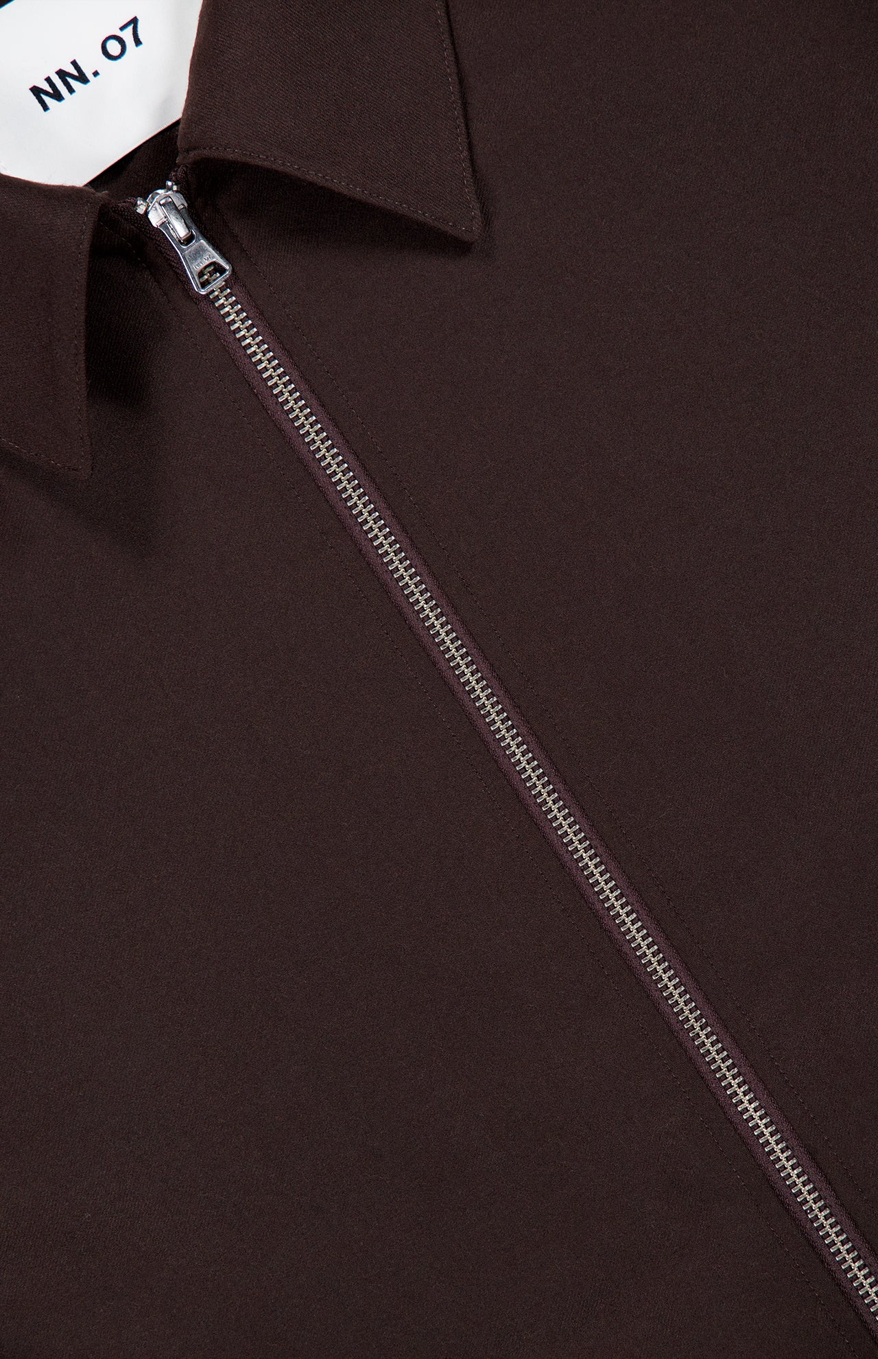 Close-up of the NN07 Alban 2-way Zip Overshirt in dark brown, featuring a pointed collar. A partial white label with “NN07” printed in black appears at the top left, emphasizing the refined details of this fall-winter overshirt-jacket crafted from responsible wool.