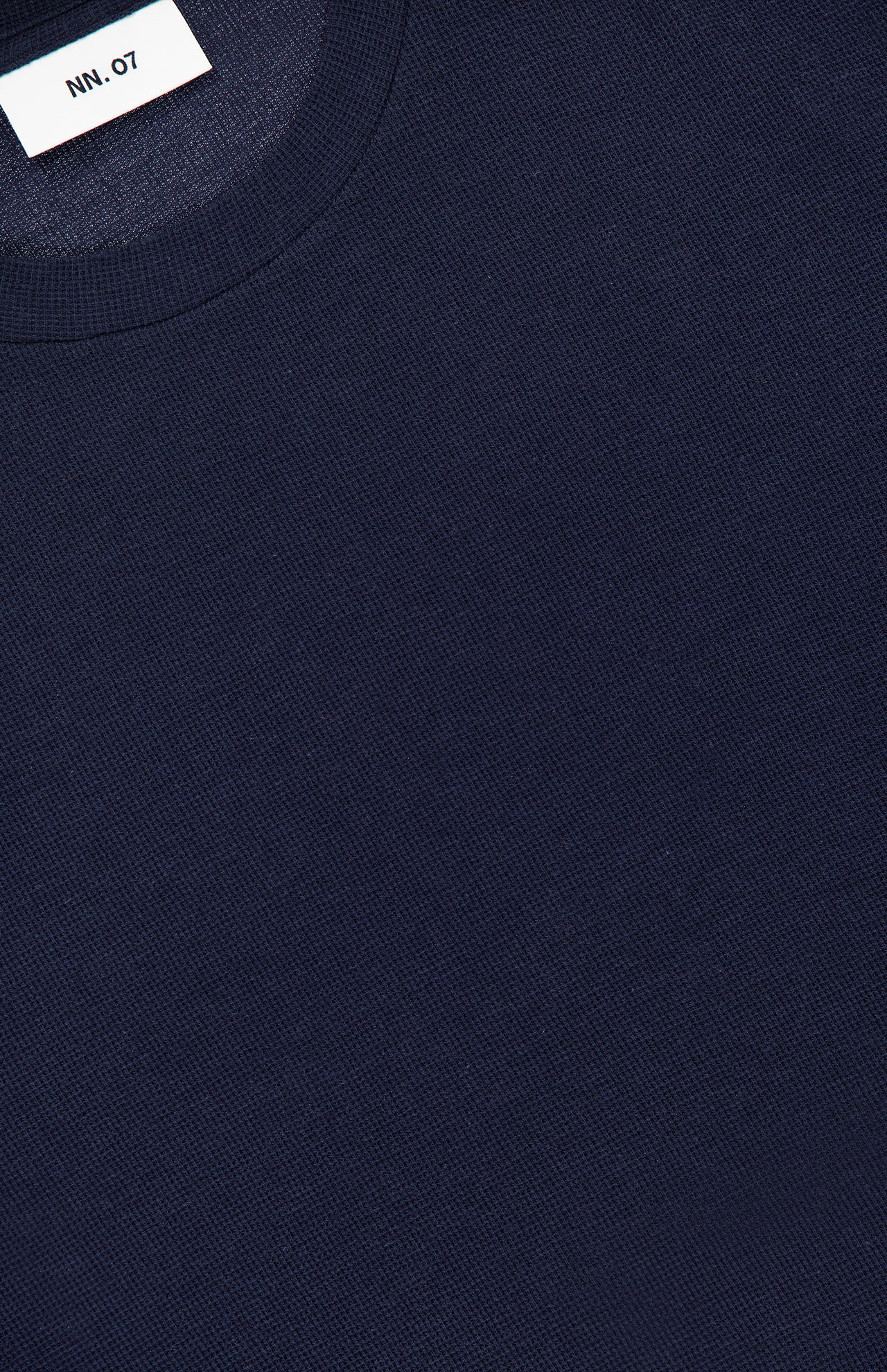 Close-up of the NN07 Clive Waffle T-Shirt in dark navy blue, featuring a round neckline and a textured waffle-knit pattern. A white label inside the collar displays the NN07 brand name in black text, adding a modern-casual touch.