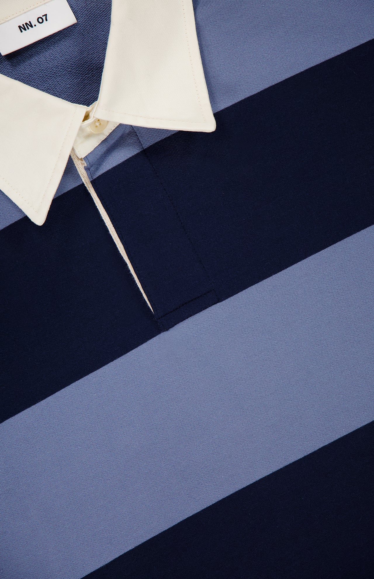 Close-up of the NN07 David Long Sleeve Polo Shirt, a sporty organic cotton shirt with wide light blue and navy stripes, white collar, branded tag, and partially unbuttoned top for a relaxed vibe.