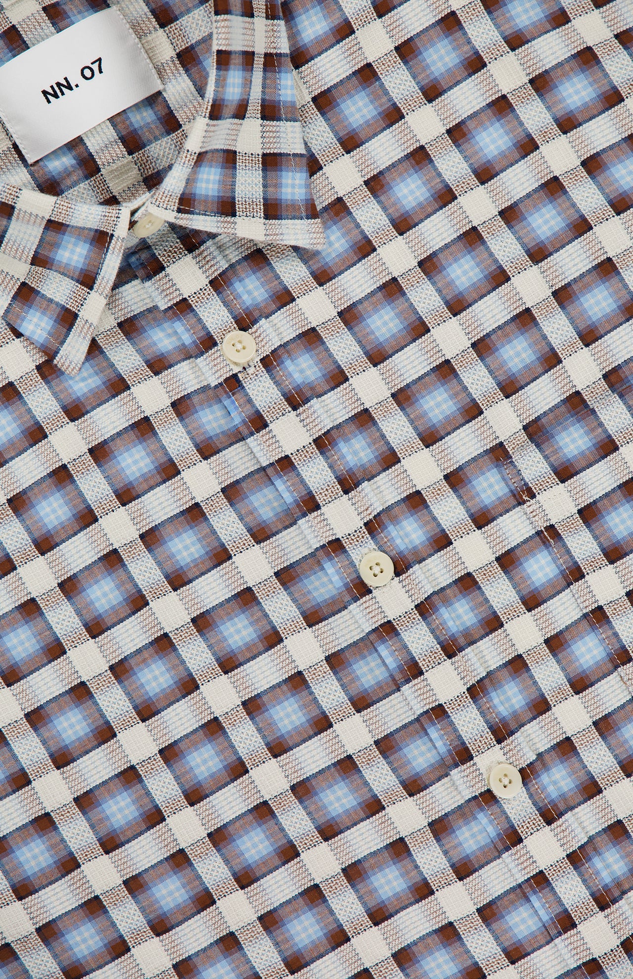 A close-up of the NN07 Deon Long Sleeve Flannel Shirt, featuring a blue, white, and brown checkered pattern in organic cotton with white buttons and a visible “NN.07” label at the neckline.