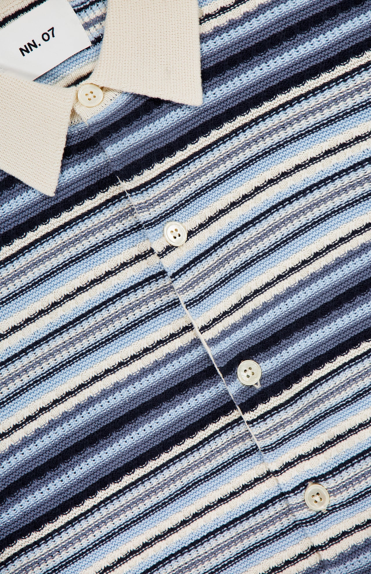 A close-up of the NN07 Nolan Polo Short Sleeve Cardigan Sweater in blue, featuring a white collar, button-down front, and navy and beige horizontal stripes. Crafted from 100% cotton with the NN07 label inside the collar.