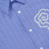 The Quinsy APP Striped Shirt by NN07 showcases light blue fabric with white, yarn-dyed stripes, classic white buttons, and a finely embroidered floral motif on the chest. The collar is finished with a label bearing the NN07 brand name.