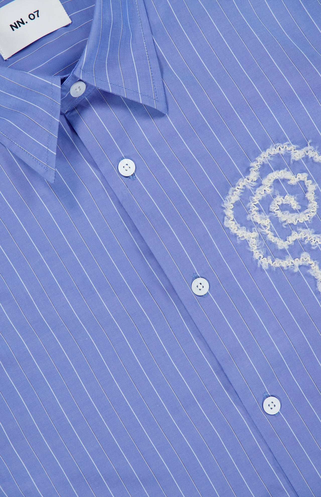 The Quinsy APP Striped Shirt by NN07 showcases light blue fabric with white, yarn-dyed stripes, classic white buttons, and a finely embroidered floral motif on the chest. The collar is finished with a label bearing the NN07 brand name.