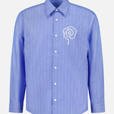 The NN07 Quinsy APP Striped Shirt is a light blue, long-sleeve button-up featuring thin, yarn-dyed white vertical stripes and a striking white embroidered spiral motif on the left chest.