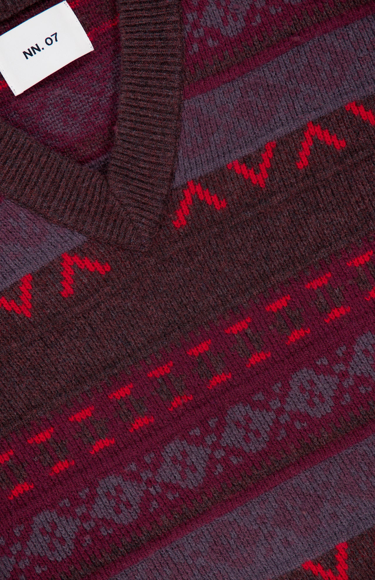 Close-up of the NN07 Tobias V-Neck Sweater, displayed in a rich berry colorway. The maroon and red patterned knit features horizontal geometric and zigzag designs, offering a modern-casual twist on the classic Fair Isle style. A white NN07 tag is visible near the collar.