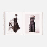 An open book, Chanel: The Impossible Collection by ASSOULINE, displays modern fashion with a woman modeling a black sheer tiered dress on the left and the same dress on a mannequin on the right—both set against luxe white minimalism.