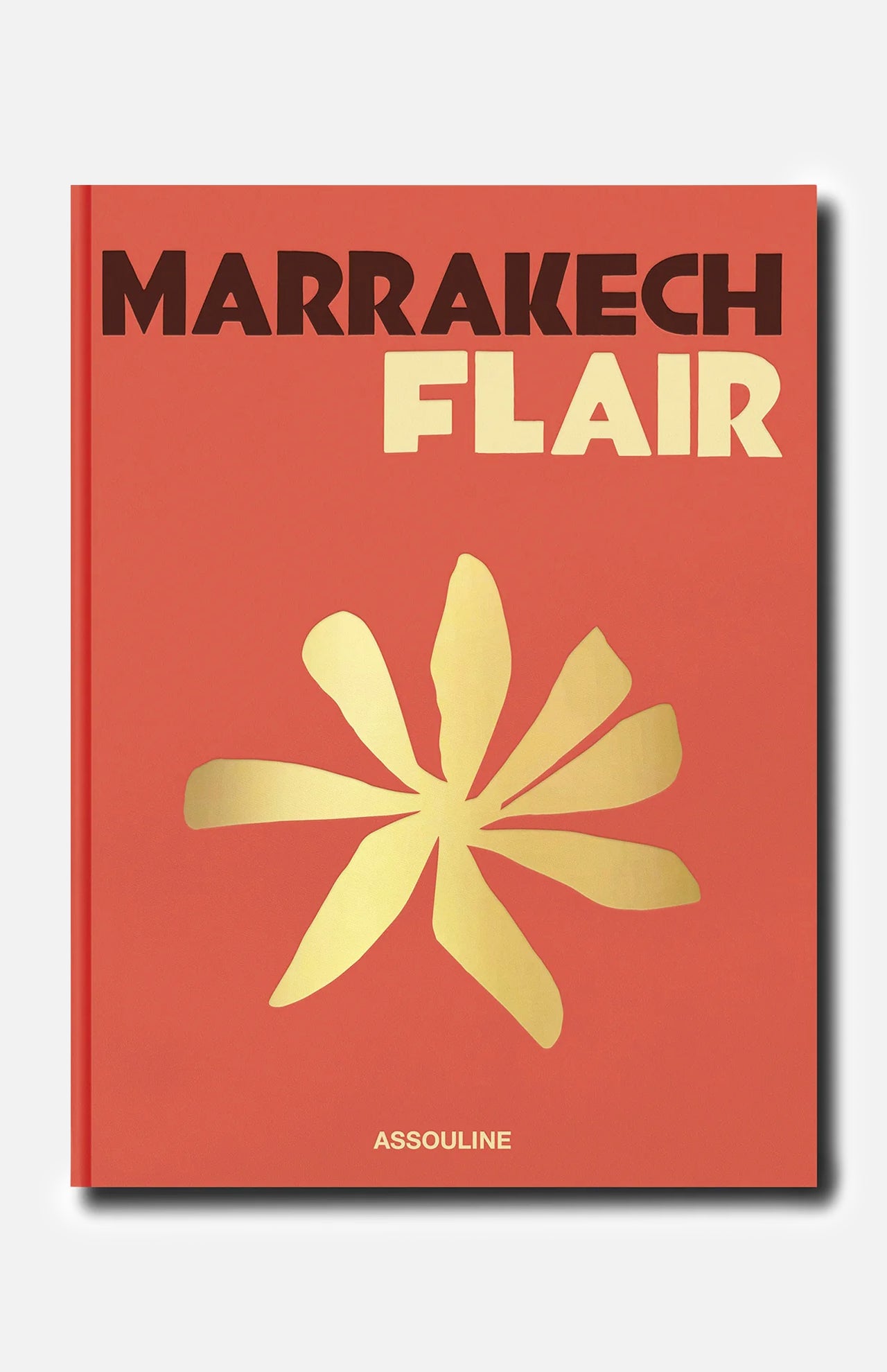 The ASSOULINE Marrakech Flair book features a striking red-orange cover inspired by the vibrant energy of the Ochre City, adorned with a bold yellow abstract flower at its center and the brand name “ASSOULINE” displayed at the bottom.