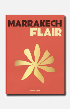 The ASSOULINE Marrakech Flair book features a striking red-orange cover inspired by the vibrant energy of the Ochre City, adorned with a bold yellow abstract flower at its center and the brand name “ASSOULINE” displayed at the bottom.