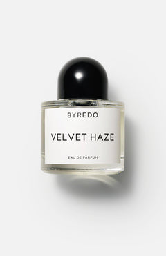 A clear glass bottle of BYREDO Velvet Haze Eau de Parfum, featuring a black round cap and minimalist white label, sits against a light background, embodying the luxury scent characteristic of BYREDO.