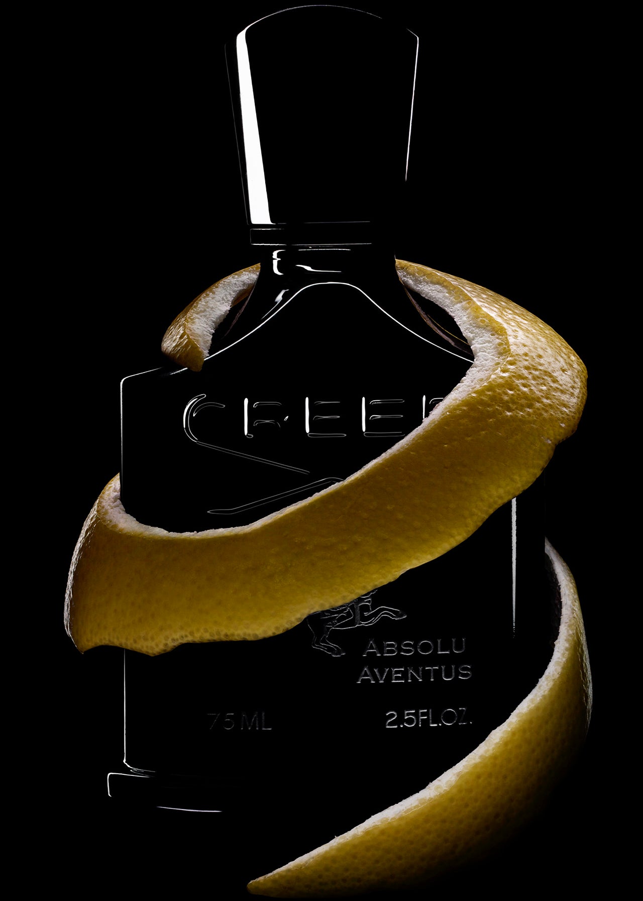 A black CREED Absolu Aventus Fragrance bottle, partly wrapped in lemon peel, stands against a dark background—its citrus scent hinted at by the 75 ML (2.5 FL.OZ.) label.