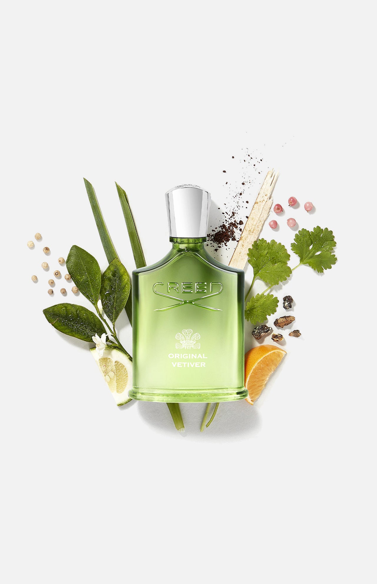 A green bottle of CREED Original Vetiver sits among citrus slices, green leaves, coriander seeds, peppercorns, and vetiver grass, emphasizing the signature fresh vetiver fragrance on a white background.