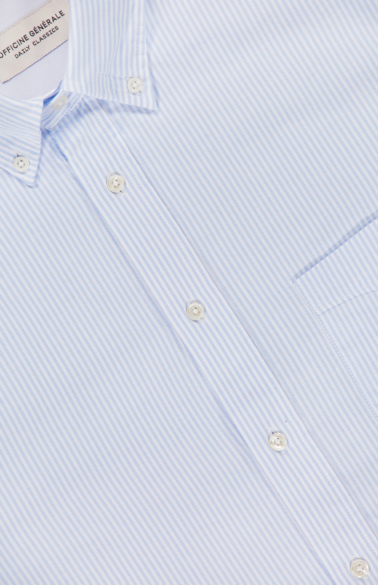Close-up of the Candy Stripe Oxford Shirt by OFFICINE GÉNÉRALE, crafted in Italian cotton with light blue and white stripes, white buttons, a left chest pocket, and a collar tag—showcasing Parisian sophistication.