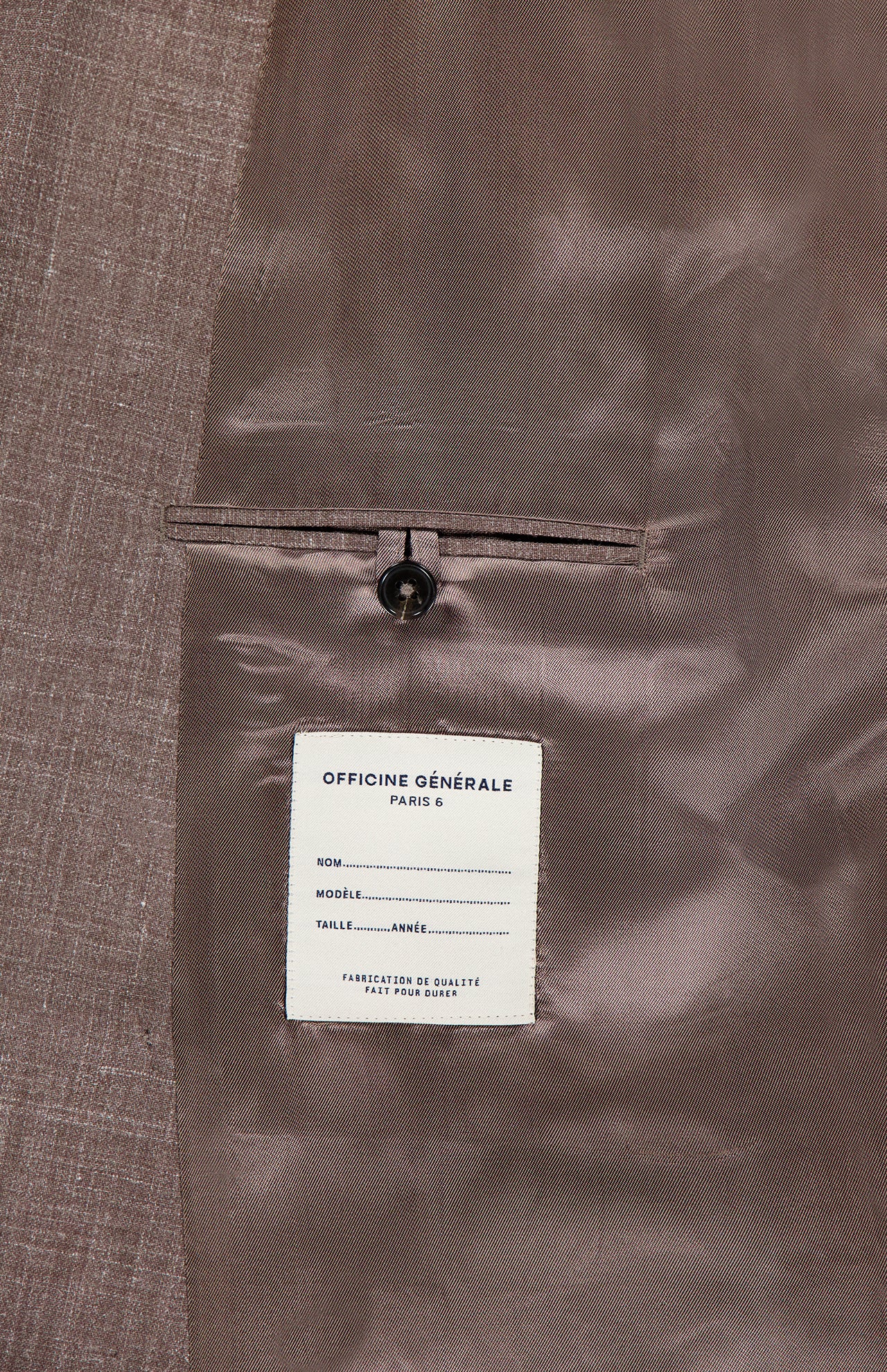 A close-up of the Leon Double Breasted Poplin Blazer by OFFICINE GÉNÉRALE shows its light brown interior, featuring a labeled inner pocket with a button. The label includes spaces for name, model, size, and year.