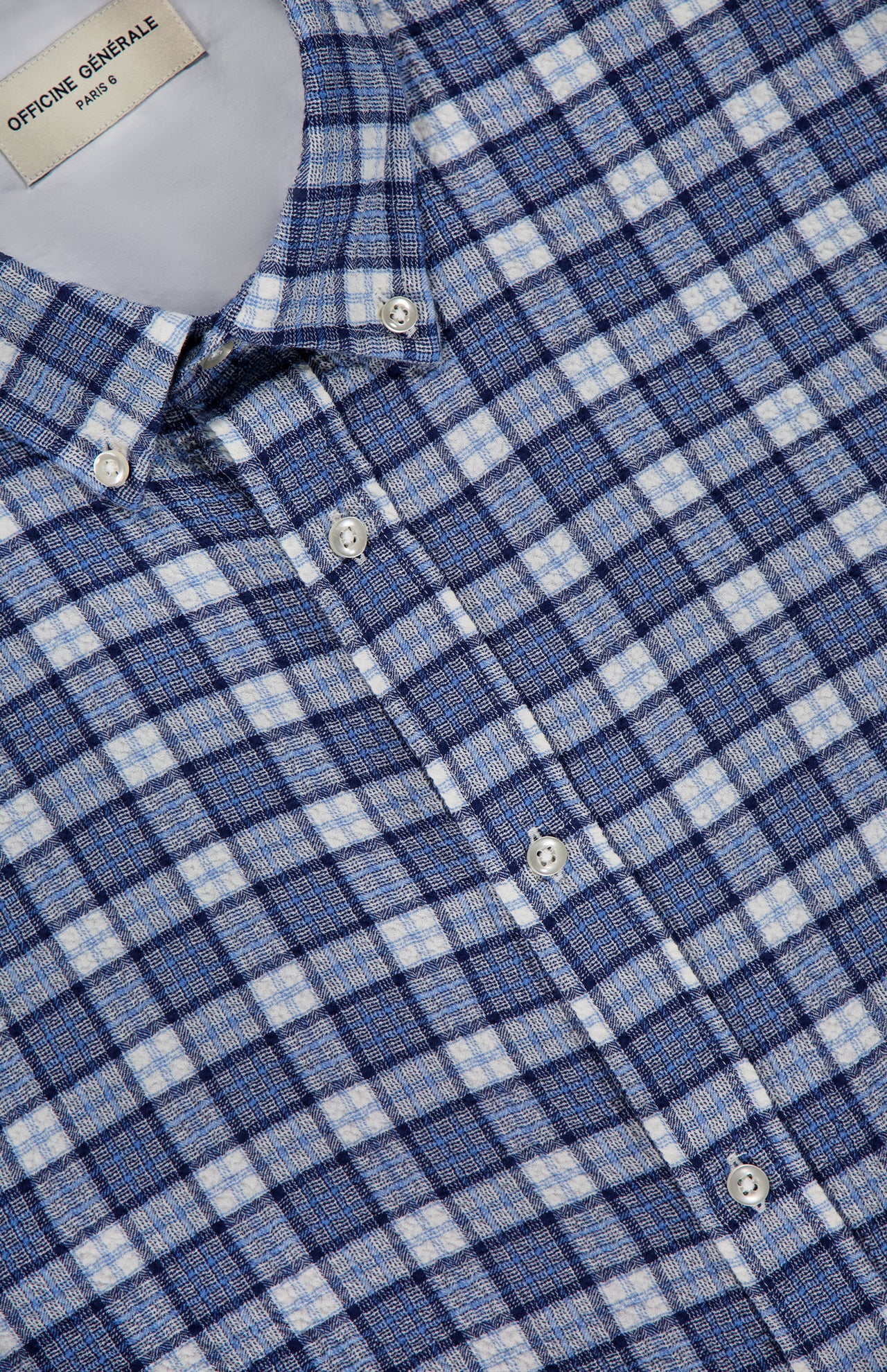 A close-up of the OFFICINE GÉNÉRALE Arsene Waffled Cotton Checkered Long Sleeve Shirt, featuring blue and white plaid with white buttons and a label inside the collar that reads OFFICINE GÉNÉRALE PARIS. This shirt is crafted from premium waffled stretch cotton for added comfort.