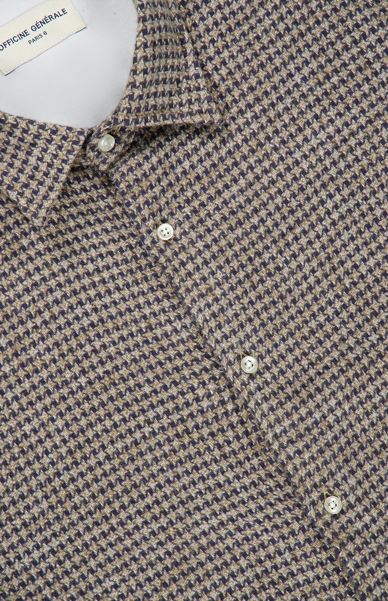 Close-up of the Giacomo Soft Cotton Houndstooth Shirt by OFFICINE GÉNÉRALE, showcasing a small collar and white buttons. The fabric displays a refined houndstooth-like texture in shades of beige, brown, and navy, capturing the timeless charm of a classic houndstooth shirt. A partially visible label at the top left hints at coordinating trouser pants available for a complete ensemble.