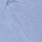 Close-up of the OFFICINE GÉNÉRALE Guido Slub Cotton Shirt in light blue, featuring a classic collar, chest pocket, subtle textured pattern, and 100% cotton fabric with a brand label at the neckline.