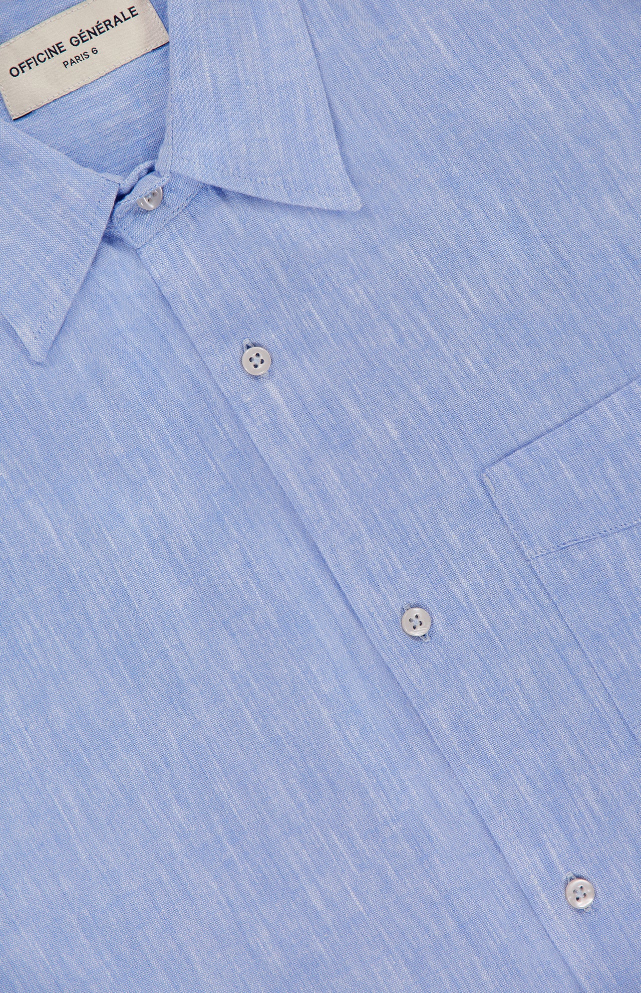 Close-up of the OFFICINE GÉNÉRALE Guido Slub Cotton Shirt in light blue, featuring a classic collar, chest pocket, subtle textured pattern, and 100% cotton fabric with a brand label at the neckline.