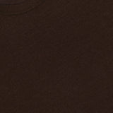 A close-up of the OFFICINE GÉNÉRALE Liev Soft Wool Long Sleeve T-Shirt in dark brown highlights its soft knit texture. A white label with partially visible text is sewn inside the collar at the top left corner.