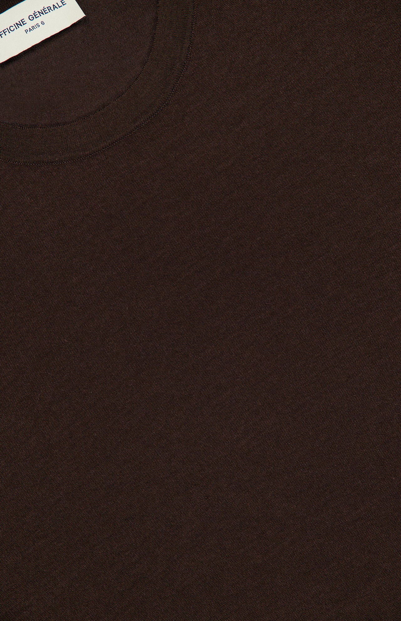 A close-up of the OFFICINE GÉNÉRALE Liev Soft Wool Long Sleeve T-Shirt in dark brown highlights its soft knit texture. A white label with partially visible text is sewn inside the collar at the top left corner.