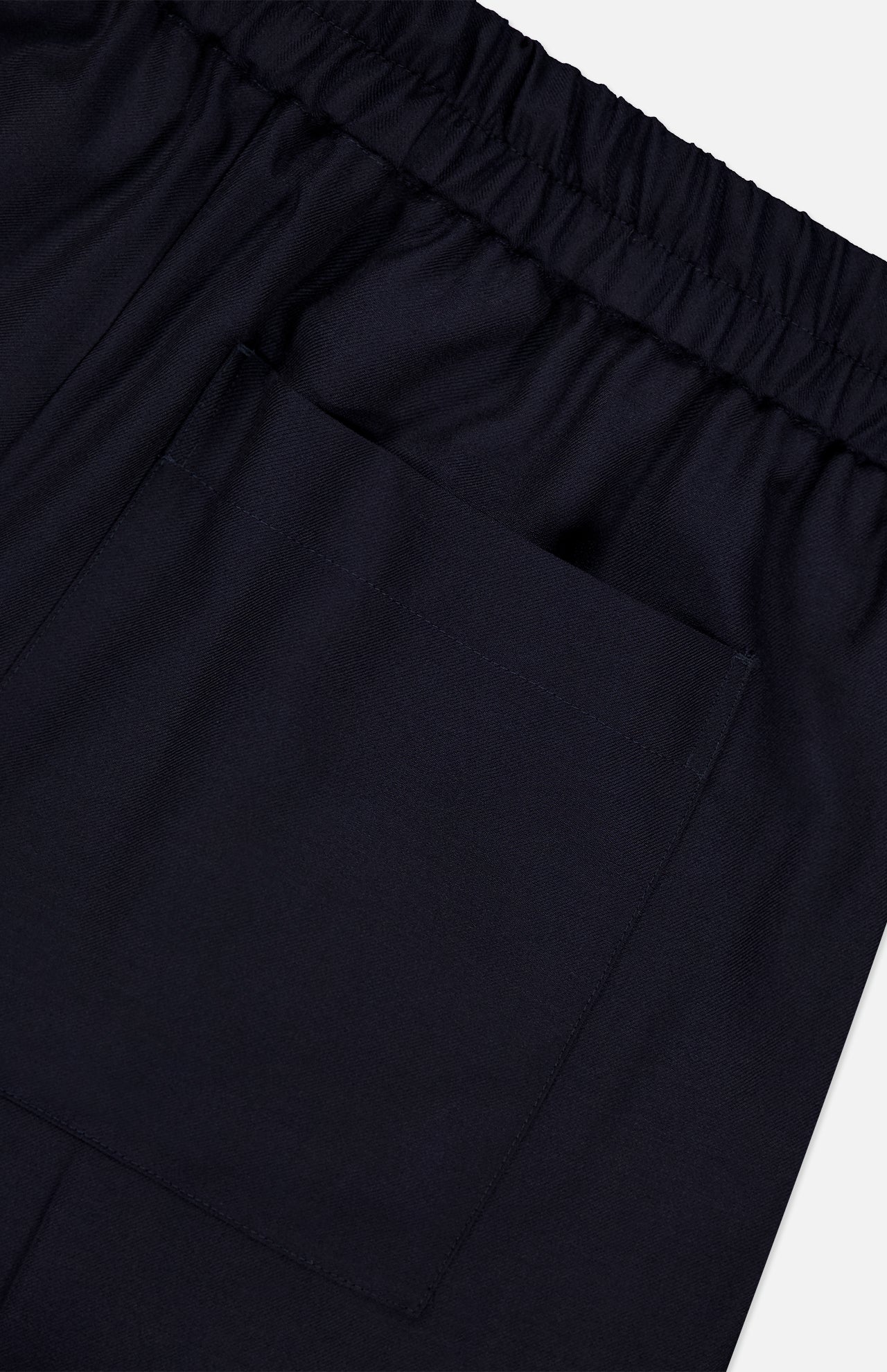 A close-up of the OFFICINE GÉNÉRALE Nelson Twill Trouser Pants in dark navy highlights the elastic waistband and a single square pocket stitched onto the smooth, slightly shiny virgin wool twill fabric.