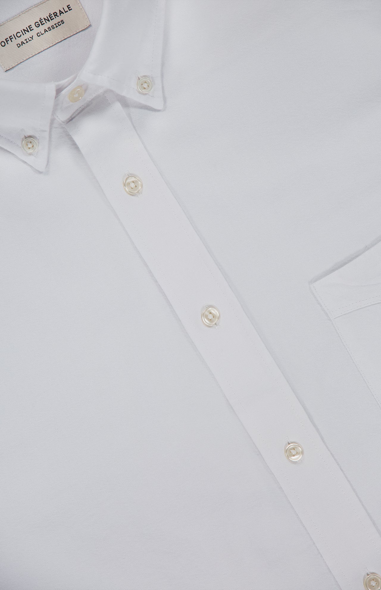 Close-up of a white oxford button-down shirt called the New Oxford Button Down Shirt by OFFICINE CREATIVE, crafted from 100% cotton, featuring a visible chest pocket and pearl-like buttons. The shirt label reads OFFICINE CREATIVE.