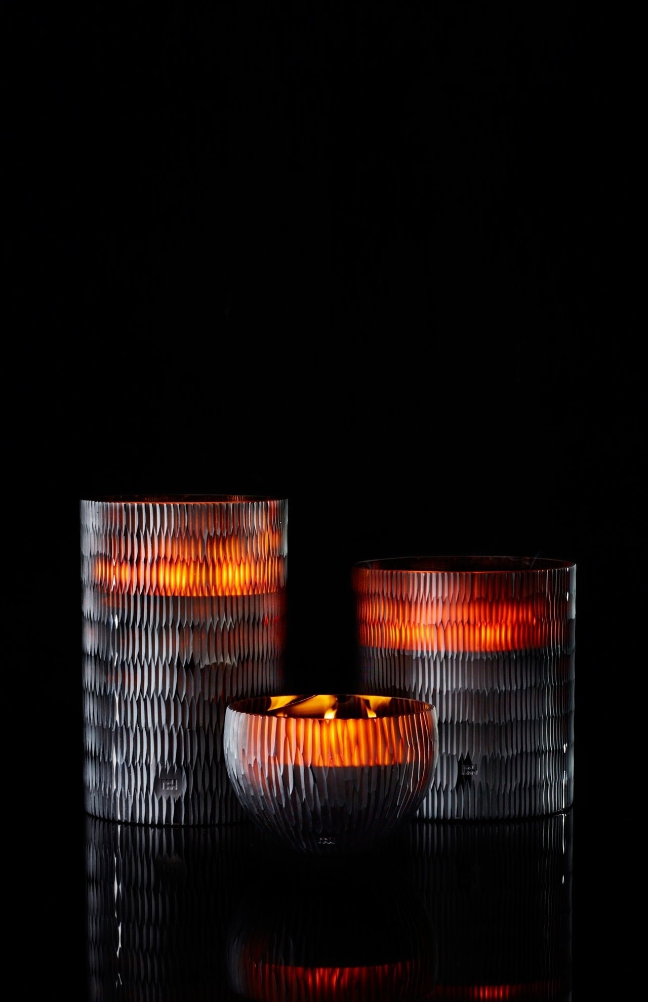 Three ONNO Amber Rhombus L Ginger Fig candle holders glow warmly against a black background. Two tall holders flank a smaller round one, creating a designer ambiance with luxury fragrance for your home.