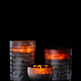 The ONNO Amber Rhombus L Sage Candle glows warmly in a textured glass holder, set against a black background, showcasing hand-poured craftsmanship and adding designer fragrance to your space.