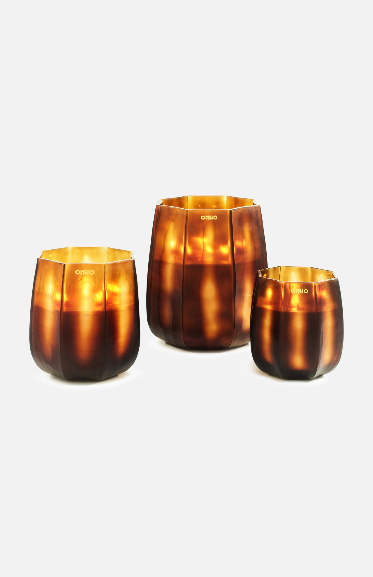Three ONNO Embrace S Sage candle holders in brown and gold glass are shown against a white background. Each features a glossy, translucent finish and gives off a warm amber glow—ideal for displaying a luxury scented candle.