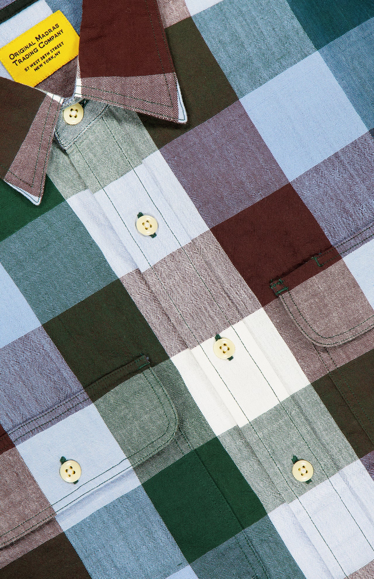 Close-up of the ORIGINAL MADRAS Check Long Sleeve Shirt, handwoven in pure Indian cotton with large green, burgundy, light blue, and white checks, yellow buttons, front pockets, and a yellow tag on the collar.