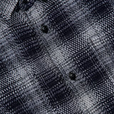 Close-up of the Check Roundtail Overshirt by ORIGINAL MADRAS, featuring a black and white plaid cotton fabric and black buttons. The yellow label with black text is partly visible in the top left corner.