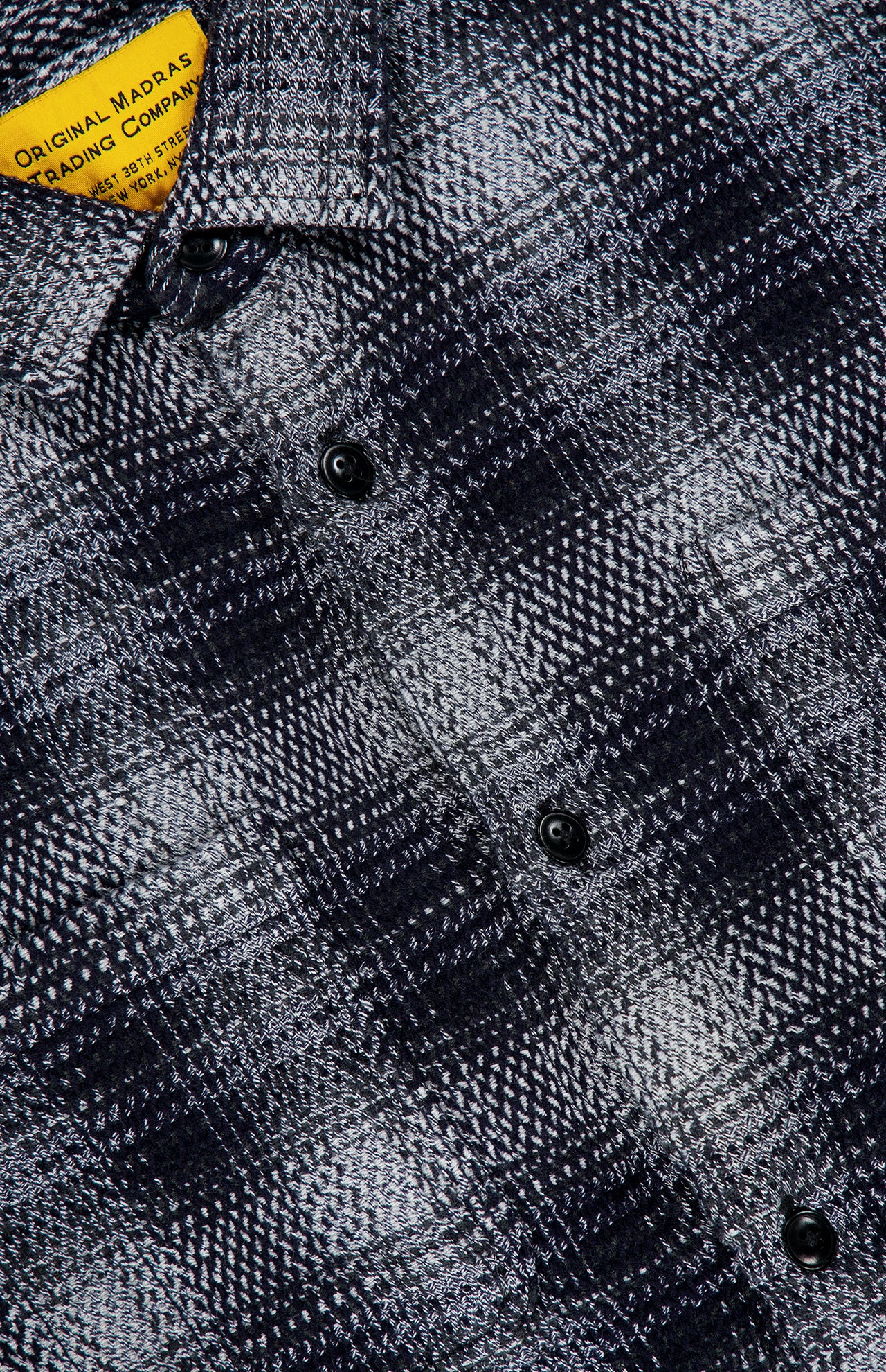 Close-up of the Check Roundtail Overshirt by ORIGINAL MADRAS, featuring a black and white plaid cotton fabric and black buttons. The yellow label with black text is partly visible in the top left corner.