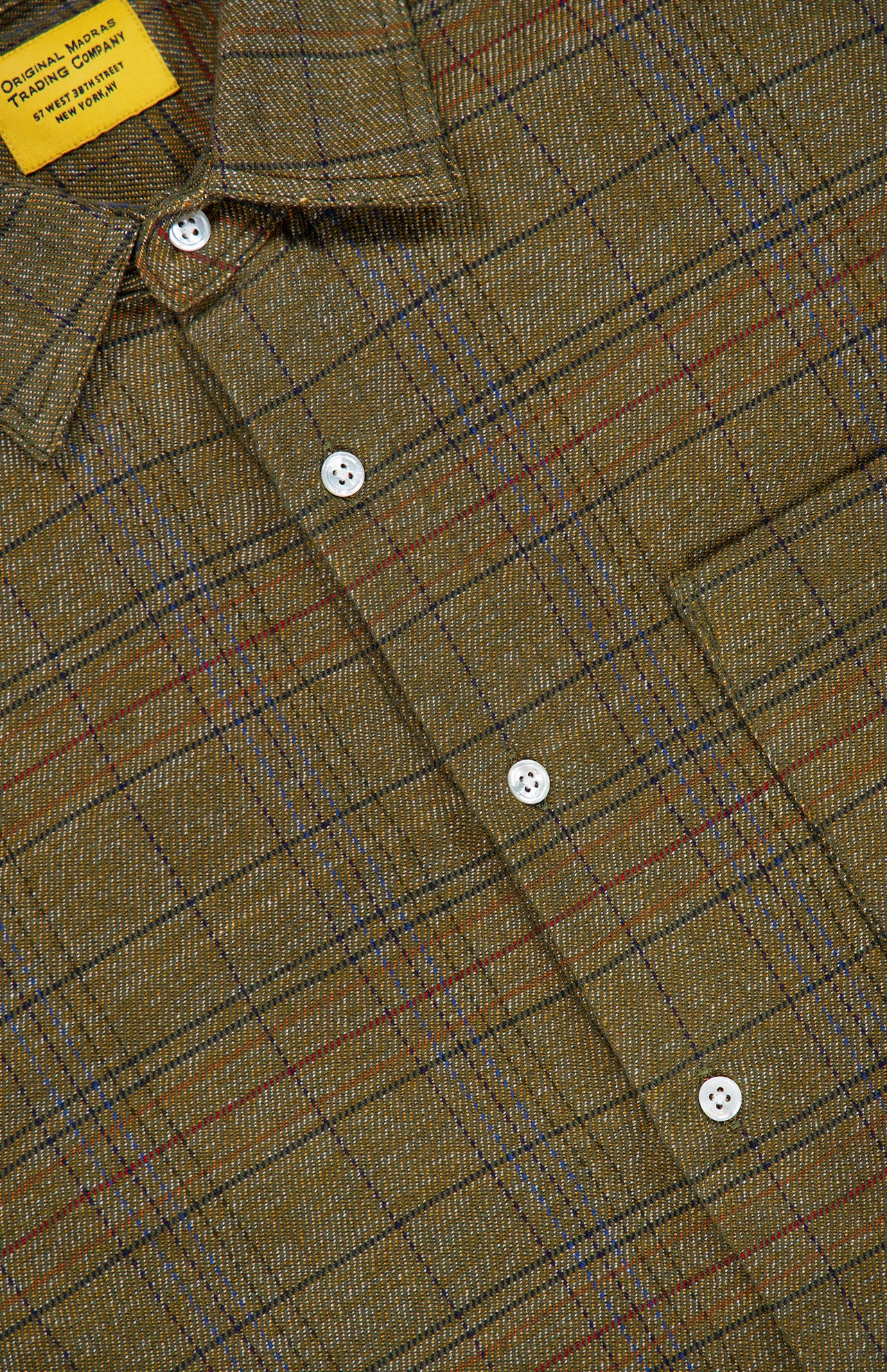 A close-up of the ORIGINAL MADRAS Check Straight Collar Dress Shirt showcases its green plaid Madras check, accented with red, blue, and brown lines in a classic checkered pattern. The white buttons and a yellow clothing label on the collar complement the shirt, which is crafted from pure Indian cotton.