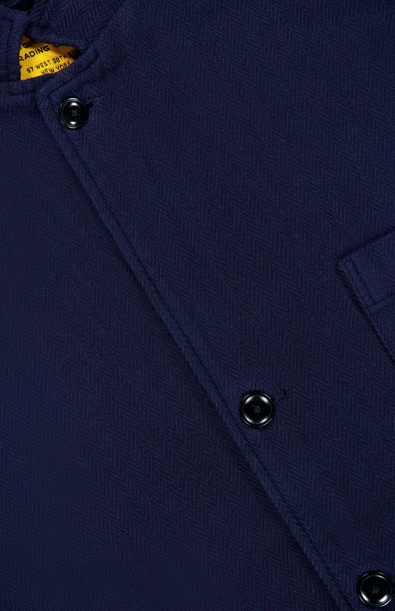 Close-up of the ORIGINAL MADRAS Herringbone Twill Soft Jacket in navy blue, featuring black buttons and a partial pocket; its refined herringbone texture stands out, while a yellow brand label is slightly visible at the top left corner.