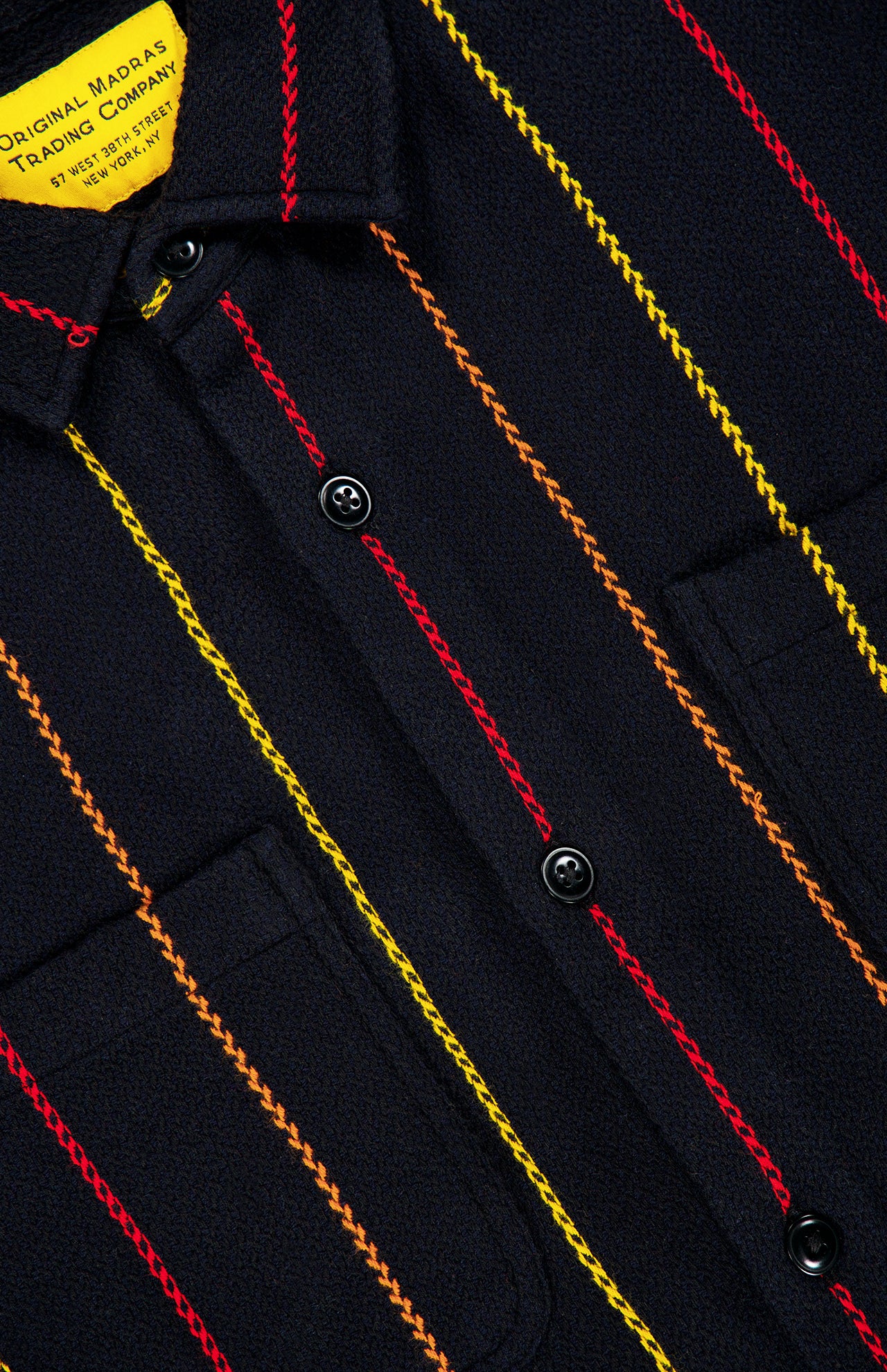 Close-up of the Pinstripe Roundtail Overshirt by ORIGINAL MADRAS, crafted from dark Indian cotton with vertical red, orange, and yellow stitched pinstripes, featuring black buttons, two chest pockets, and a yellow label inside the collar.