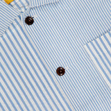 Close-up of the ORIGINAL MADRAS Seersucker Stripe Shirt Jacket, featuring blue and white stripes, brown buttons, a right chest pocket, and a yellow “Original Madras Trading Company” label.