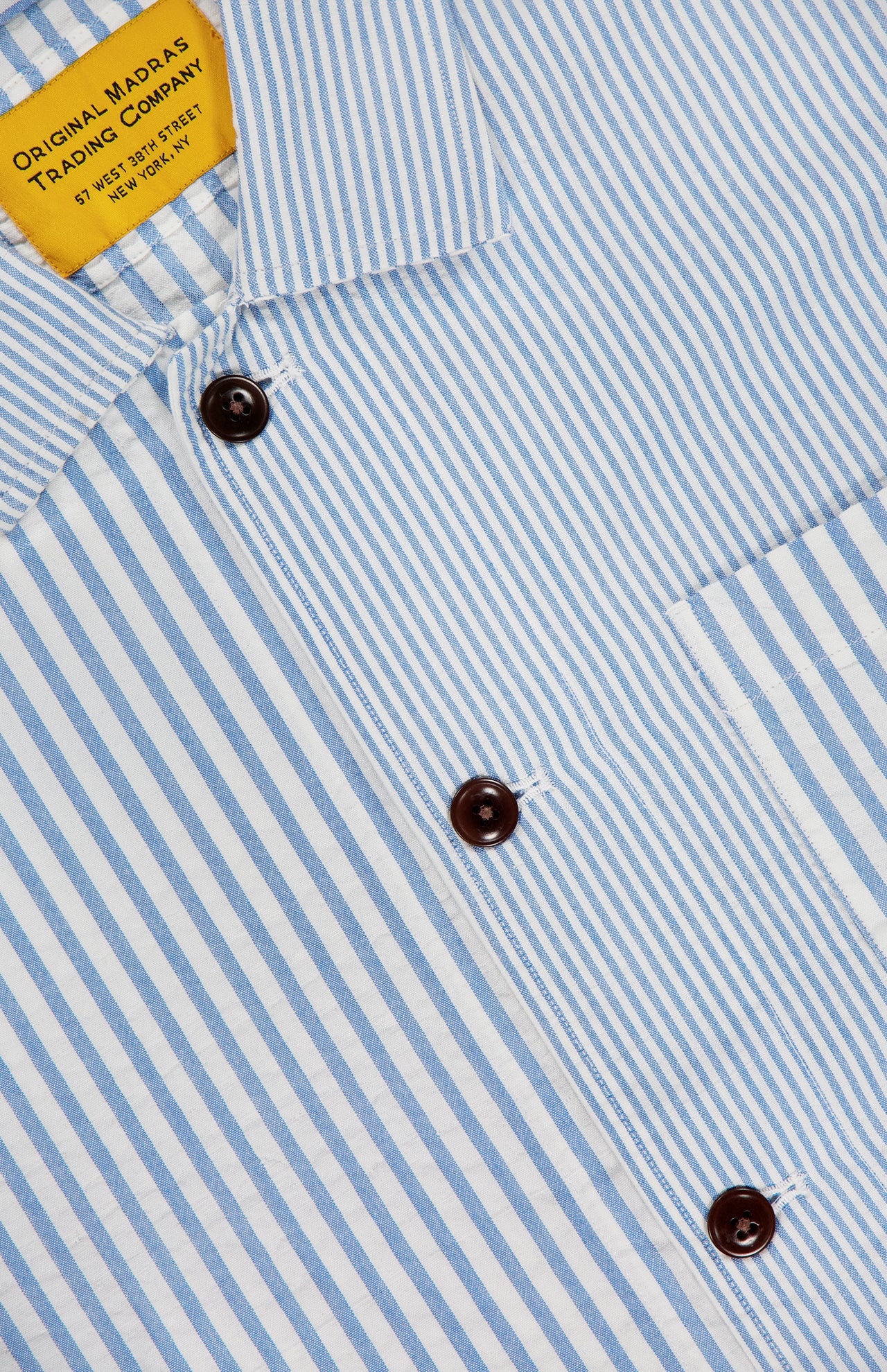 Close-up of the ORIGINAL MADRAS Seersucker Stripe Shirt Jacket, featuring blue and white stripes, brown buttons, a right chest pocket, and a yellow “Original Madras Trading Company” label.