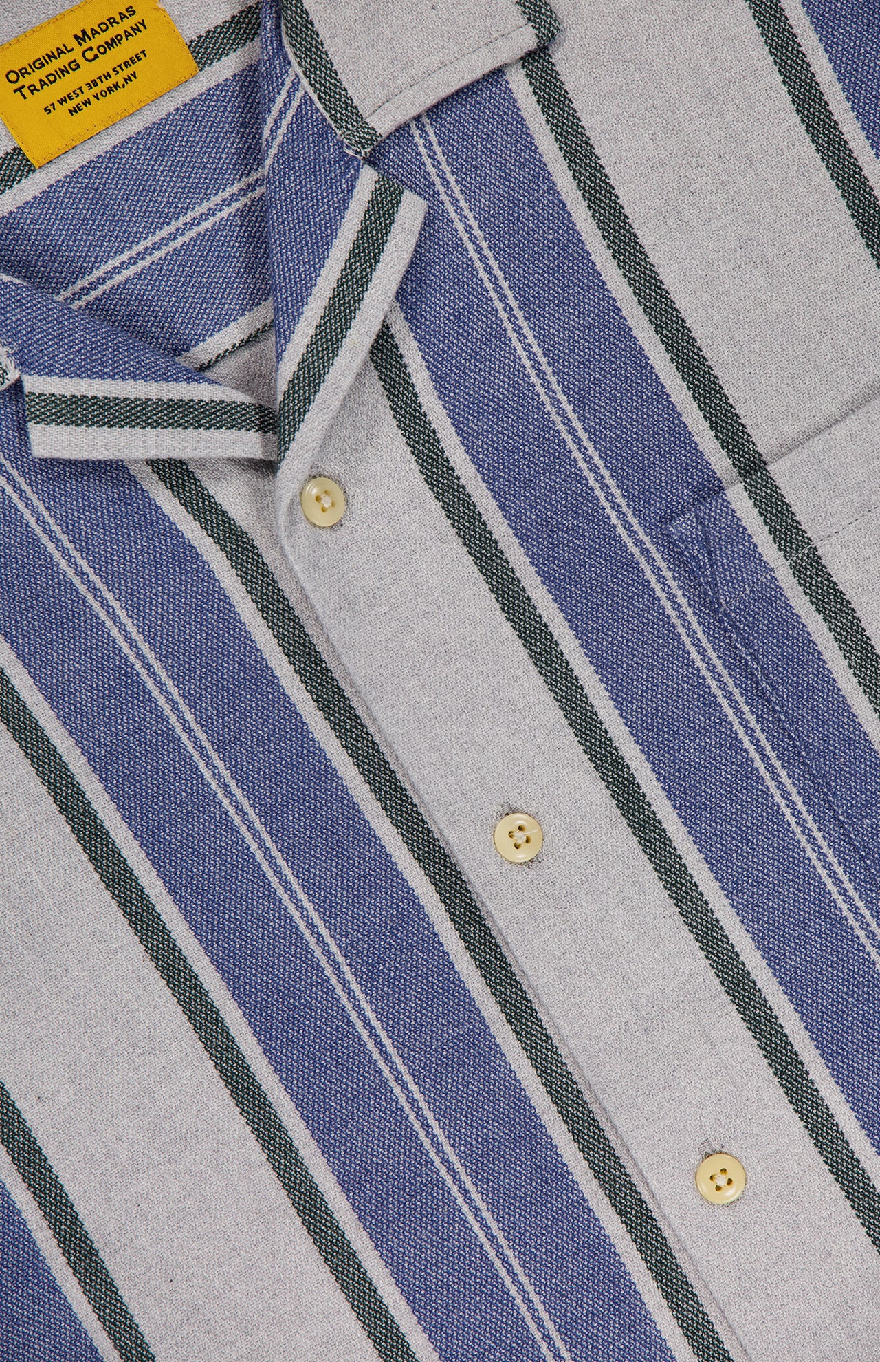 Close-up of the ORIGINAL MADRAS Stripe Lax Camp Shirt made from pure Indian cotton with vertical blue, green, and white stripes, beige buttons, and a yellow neck tag reading Original Made Trading Company. Fabric has a textured look.