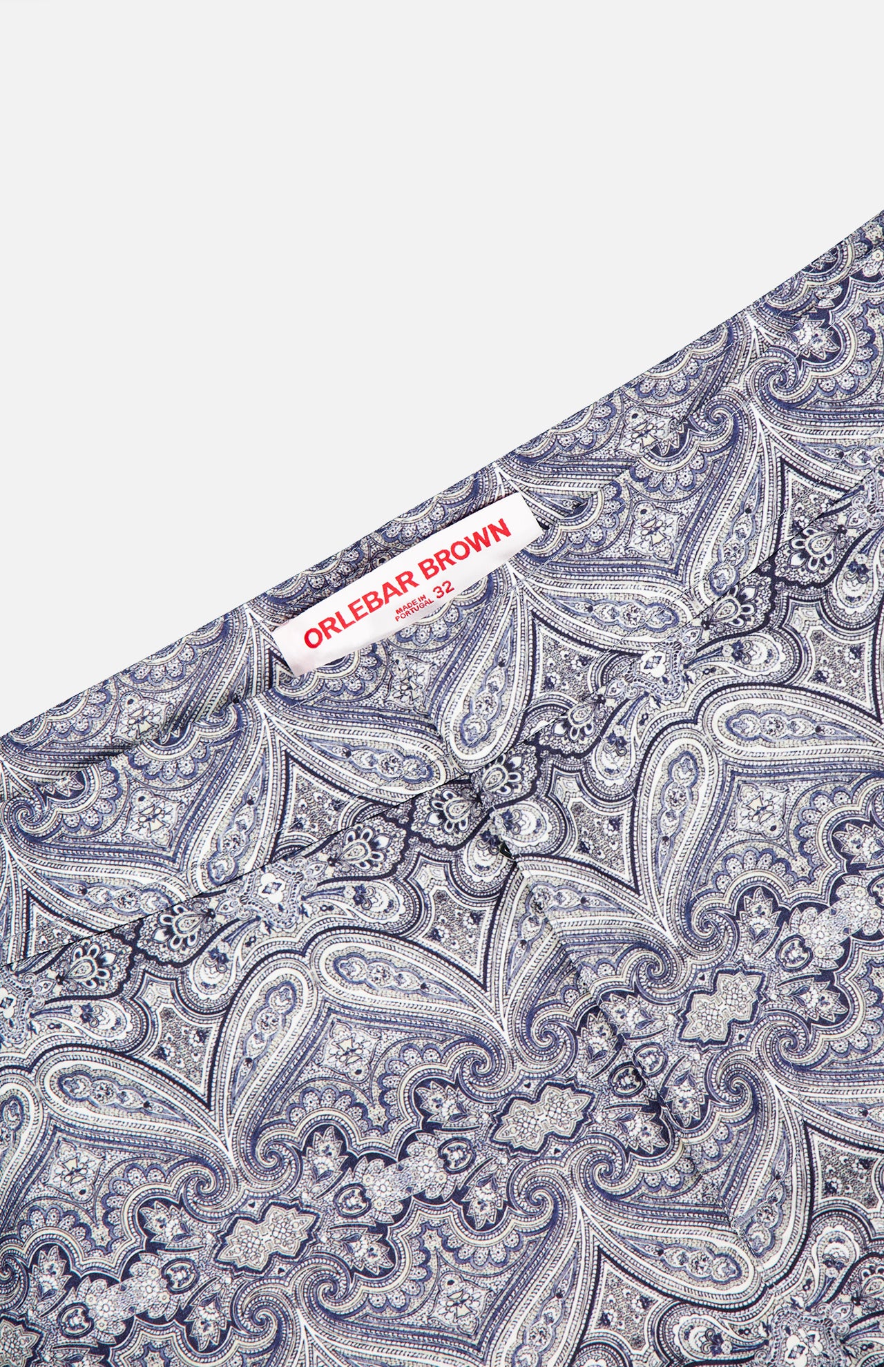 Close-up of ORLEBAR BROWN Bulldog Carnival Swim Trunk in blue and white paisley print, featuring “ORLEBAR BROWN” and “32” on the sewn label, adjustable side buckles, and extra branding details in small print below.