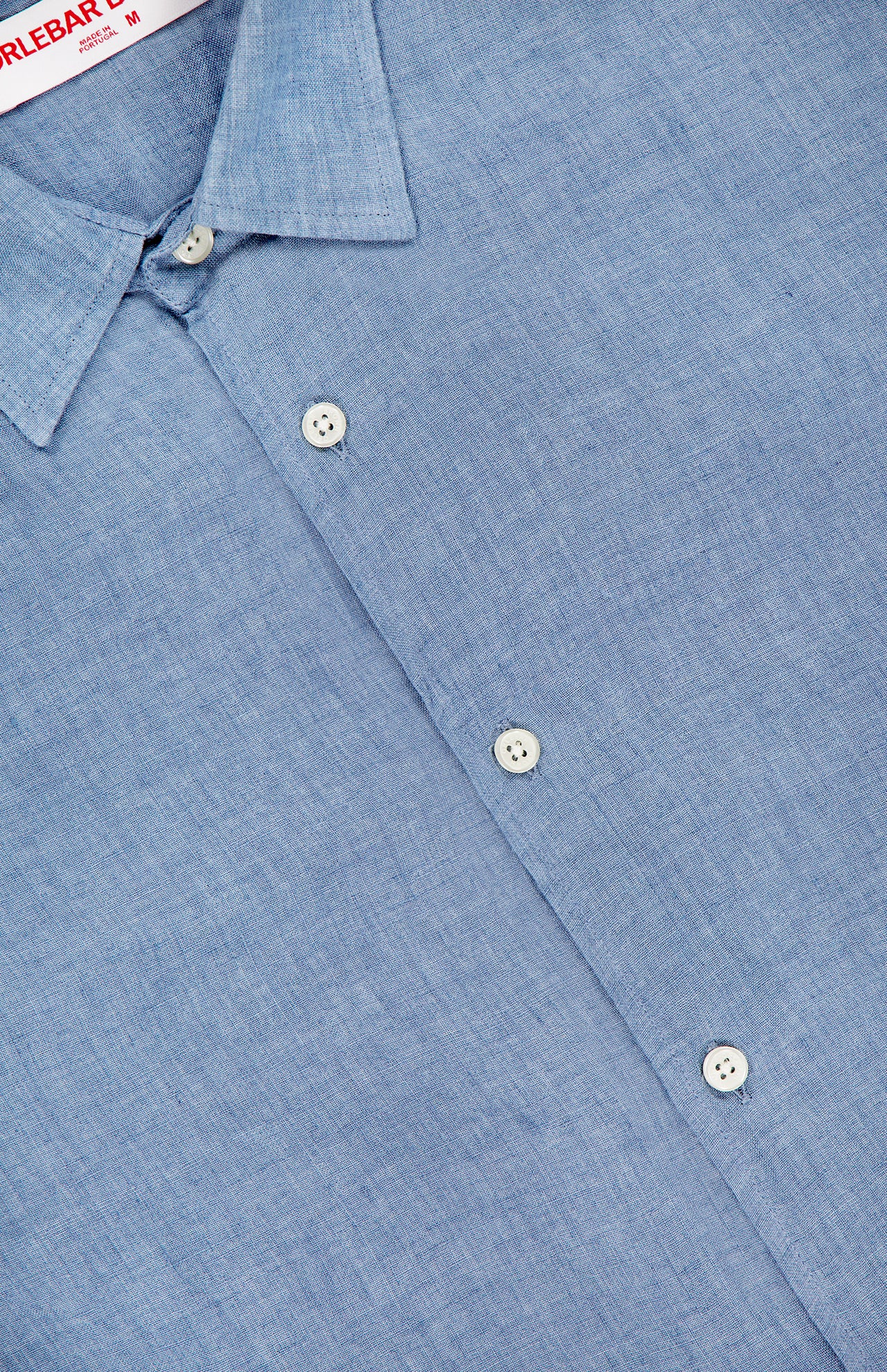 A close-up of the ORLEBAR BROWN Giles Lino Stitched Shirt in light blue linen features white buttons, a pointed collar, and a partially visible red and white ORLEBAR BROWN label at the top.