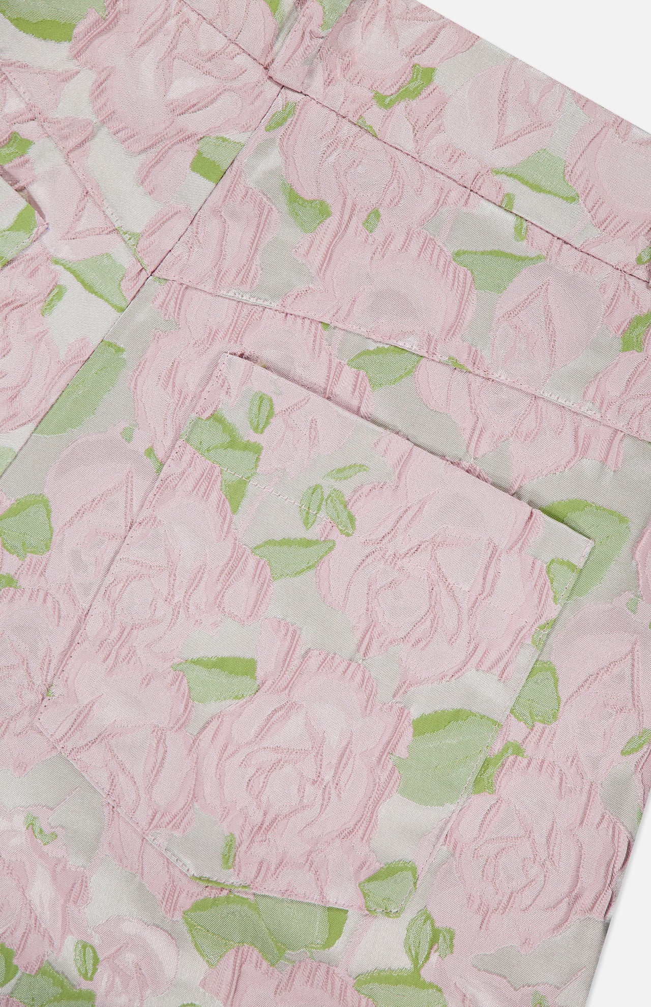 Close-up of fabric from PATBO's Garden Jacquard Pants, showcasing a delicate pink floral pattern with green leaves that evokes a garden party feel. The image highlights a pocket sewn onto the textured, woven material, emphasizing the intricate flower motif and pastel jacquard design.
