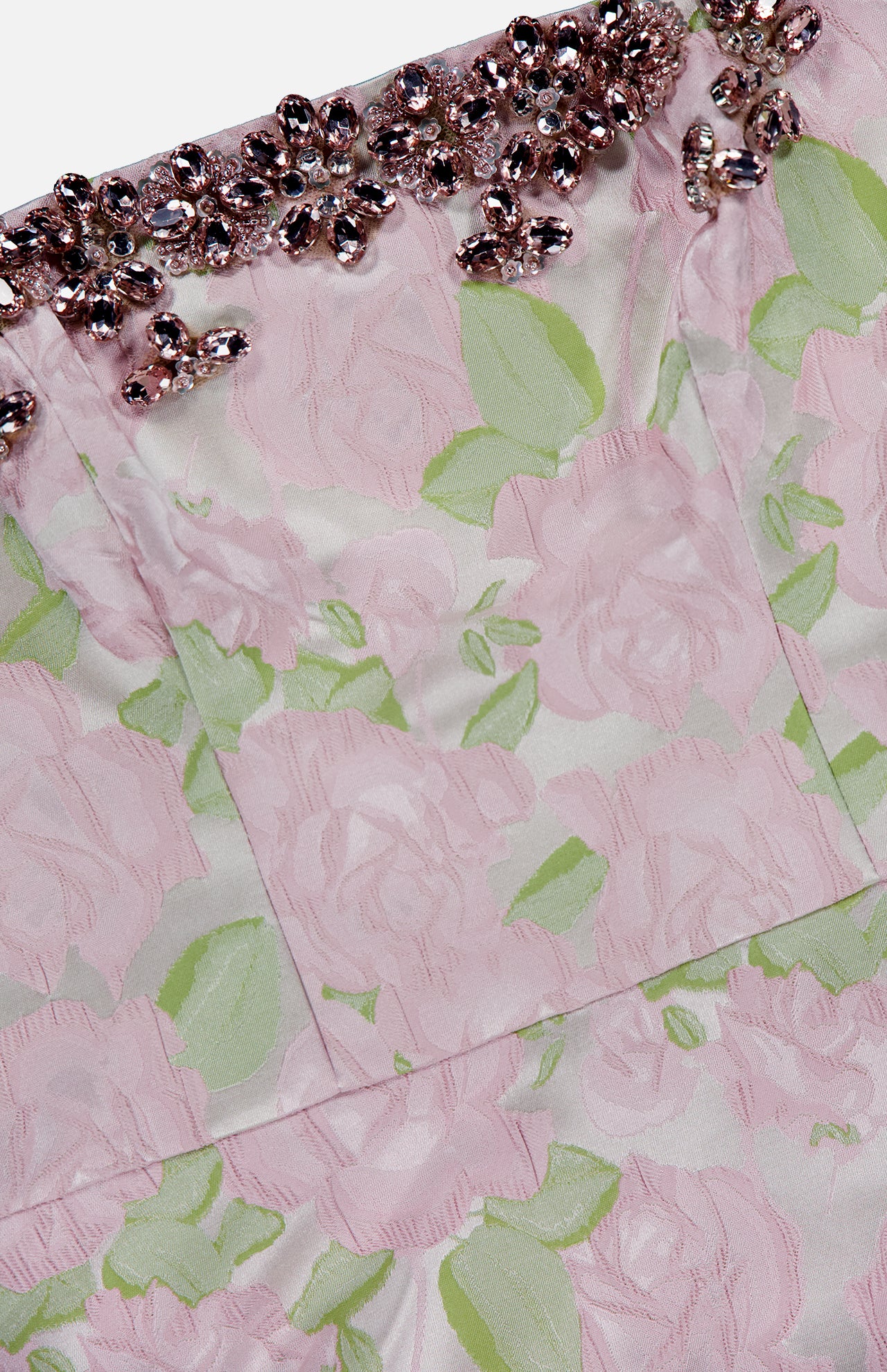 A close-up of the PATBO Garden Jacquard Crystal Top reveals a delicate pink floral jacquard fabric adorned with green leaves and accented by an embellished trim of pink and silver gemstones along the top edge. The textured rose pattern exudes pastel elegance reminiscent of a corset top, while the sparkling crystal details create a striking, statement look ideal for nights out.