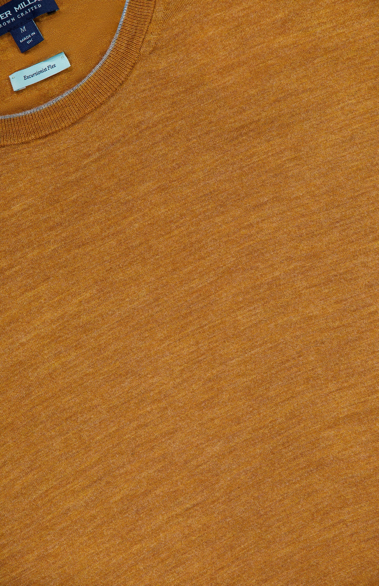 Close-up of the Excursionist Flex Crewneck Sweater in an orange-brown hue, highlighting the collar detail and a tag displaying the PETER MILLAR brand and size information. The fabric looks soft with a subtly heathered finish, evoking the comfort of merino wool—ideal for travel or everyday wear.