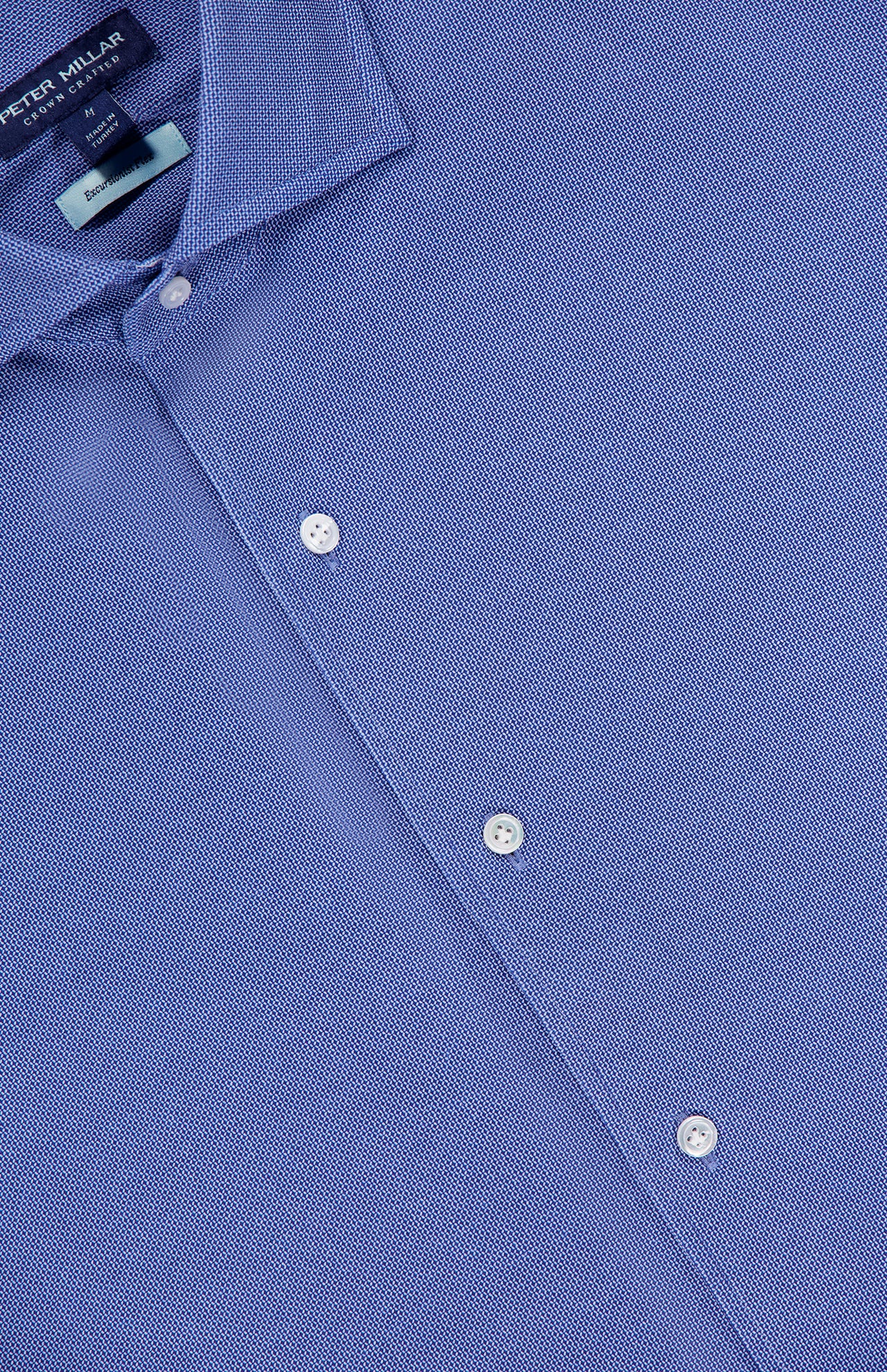 Close-up of the Excursionist Flex Print Sport Shirt in blue, crafted from cotton-stretch fabric and featuring white buttons. The shirt’s label at the collar displays the PETER MILLAR brand. The wrinkle-resistant material appears smooth and neatly pressed.