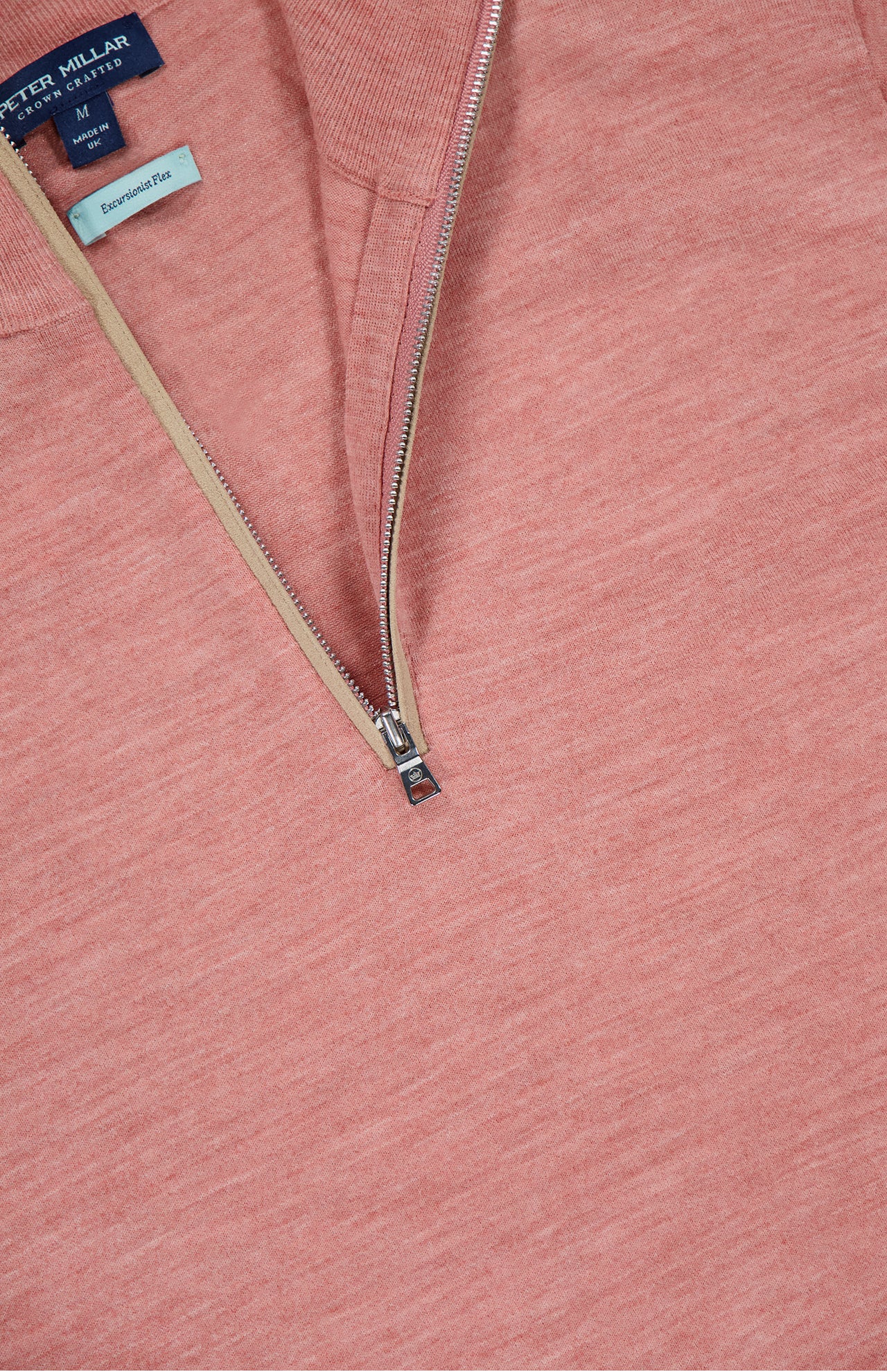 A close-up of the PETER MILLAR Excursionist Flex Quarter-Zip Sweater in salmon pink with a metal zipper and beige lining. Tags display “PETER MILLAR,” “M,” “Essentials,” and “Classic Fit”—ideal as a lightweight travel sweater.