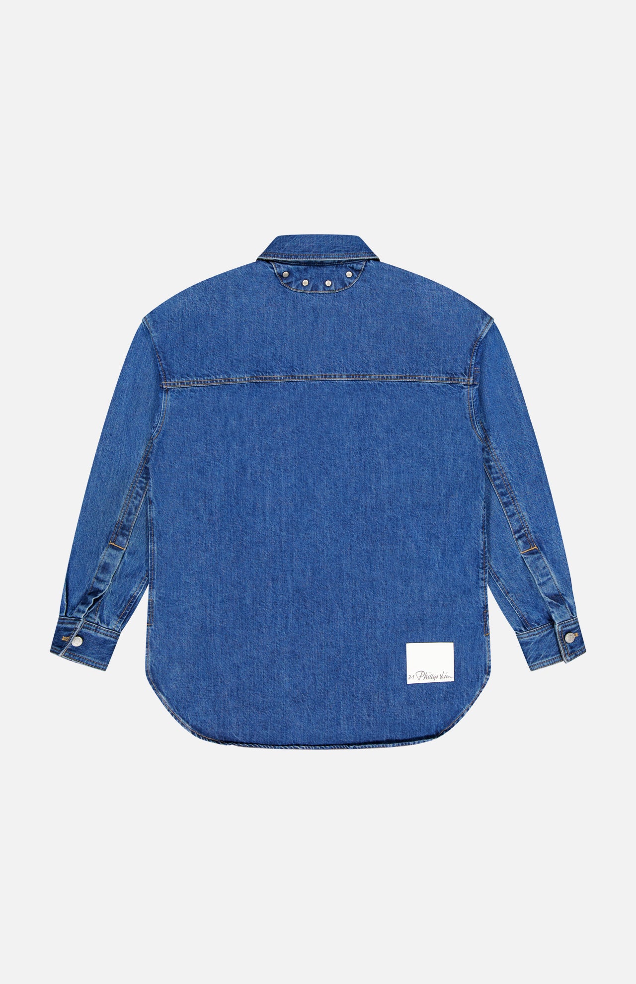 The Oversized Denim Shirt Jacket by 3.1 PHILLIP LIM is a long-sleeved blue jean shirt with a collar, silver cuff buttons, silver studs near the collar, and a white rectangular patch above the bottom hem, shown from the back.
