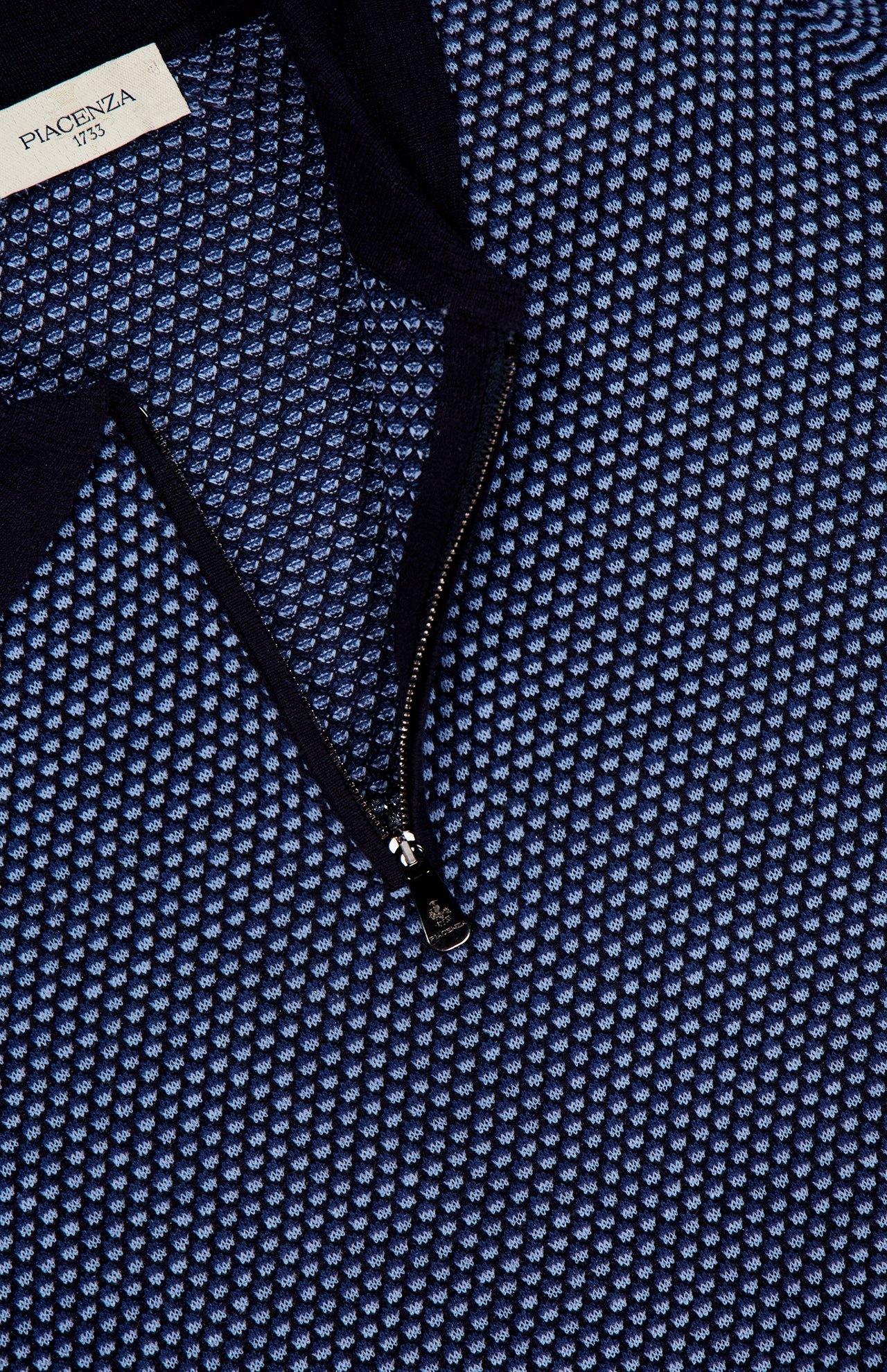 Close-up of a textured blue and black PIACENZA 1/4 Zip Knit Polo Shirt with a small repeating pattern. A label at the collar signals designer quality, while the zipper reveals more of its intricate knit.