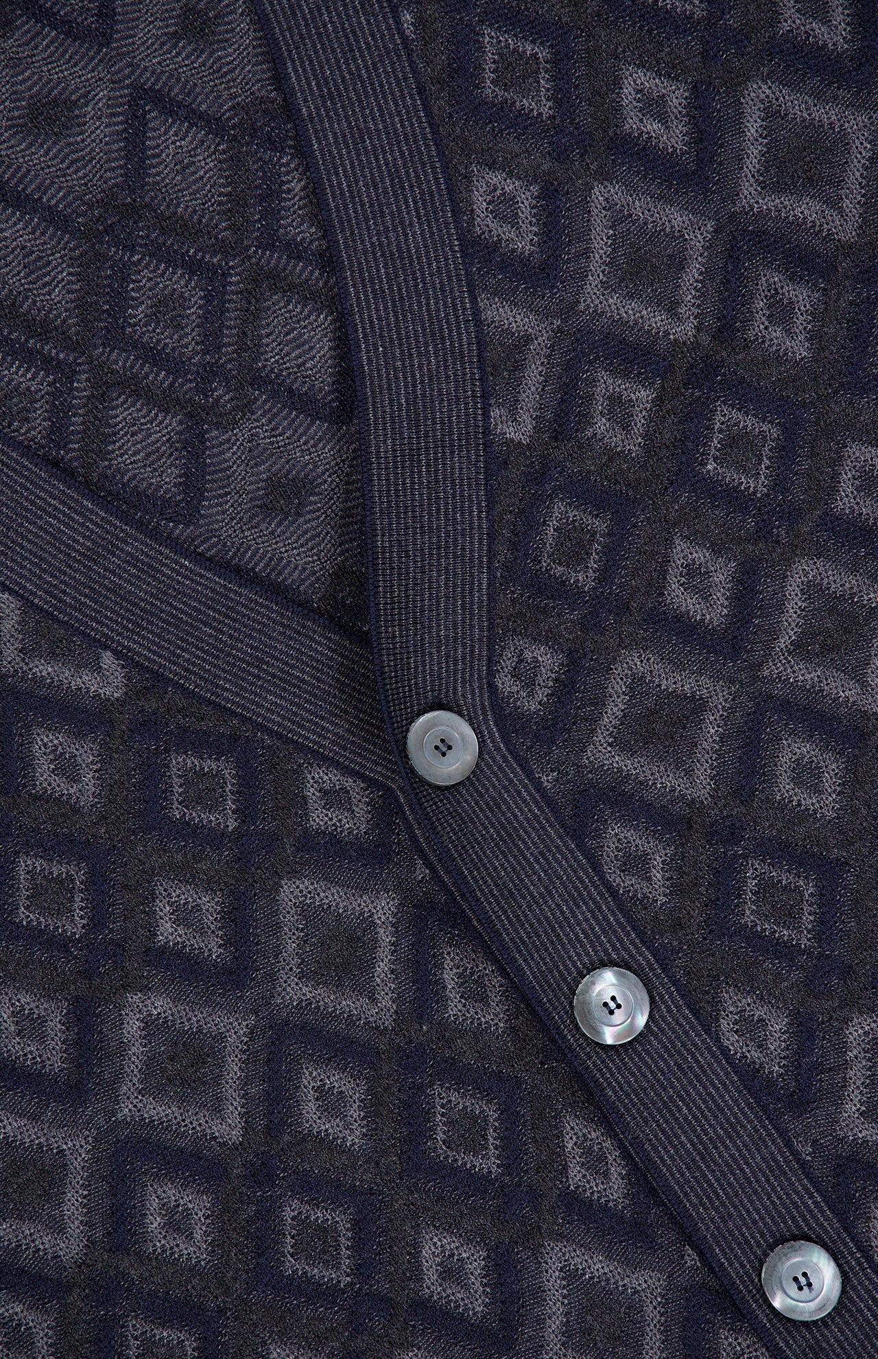Close-up of the PIACENZA Icon Argyle Stitch Cardigan, featuring a dark geometric pattern. A ribbed band with three round, silver buttons runs diagonally across the fabric.