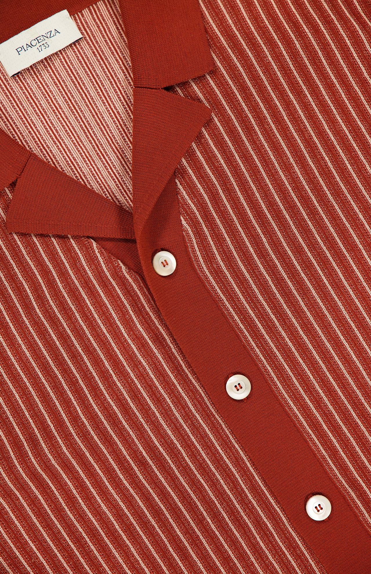 A close-up of the PIACENZA Striped Knit Short Sleeve Button Up Polo Shirt, featuring rust color, white vertical pinstripes, and white buttons, showcases Italian craftsmanship and the signature PIACENZA tag.