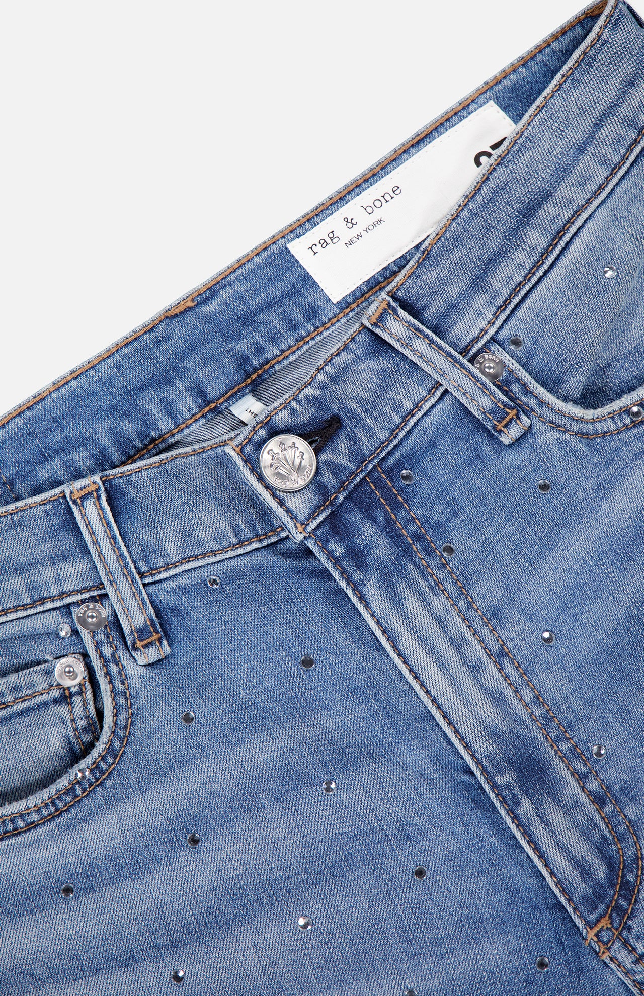 Close-up of RAG & BONE Harlow Ankle Straight Jean in light blue denim with silver studs, front pockets, belt loops, a metal button, and a white brand label on the waistband; classic straight leg design.