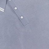 A close-up of the RAG & BONE Harvey Tipped Polo Shirt in light grey shows white buttons and a ribbed collar with two thin white stripes, plus the partially visible shirt tag at the neckline.